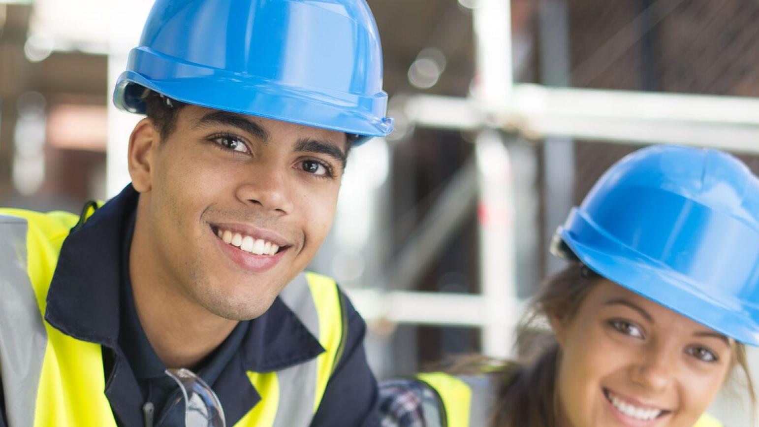 Junger Mann und junge Frau mit blauem Helm auf einer Baustelle lächeln in die Kamera