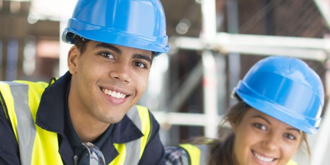 Junger Mann und junge Frau mit blauem Helm auf einer Baustelle lächeln in die Kamera