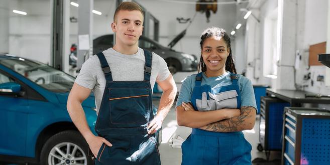 Zwei Jugendliche stehen in einer KFZ-Werkstatt und lächeln in die Kamera.