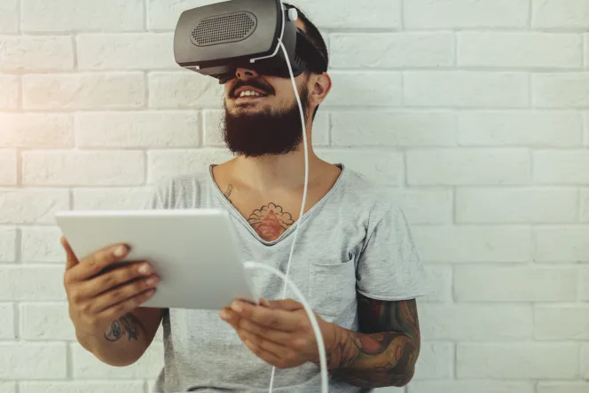 ein junger Mann hält ein Tablet in der Hand und trägt eine VR-Brille.