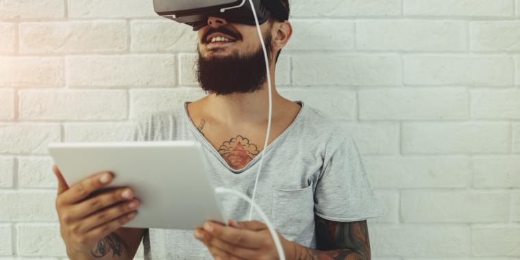 ein junger Mann hält ein Tablet in der Hand und trägt eine VR-Brille.
