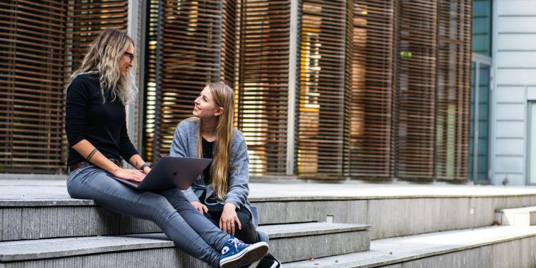 zwei Frauen sitzen mit einem Laptop vor einem Universitätsgebäude