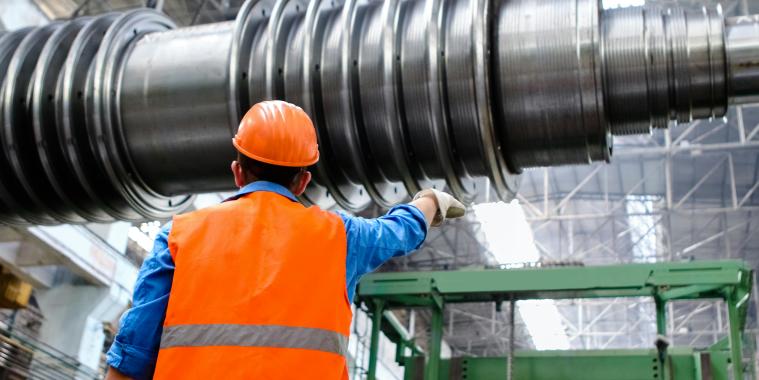 Ein Mann mit Helm und Warnweste deutet auf ein großes Metallteil in einer großen Industrielagerhalle
