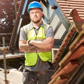 Ein Handwerker steht mit Warnweste, blauem Helm und verschränkten Armen vor einem Hausdach.